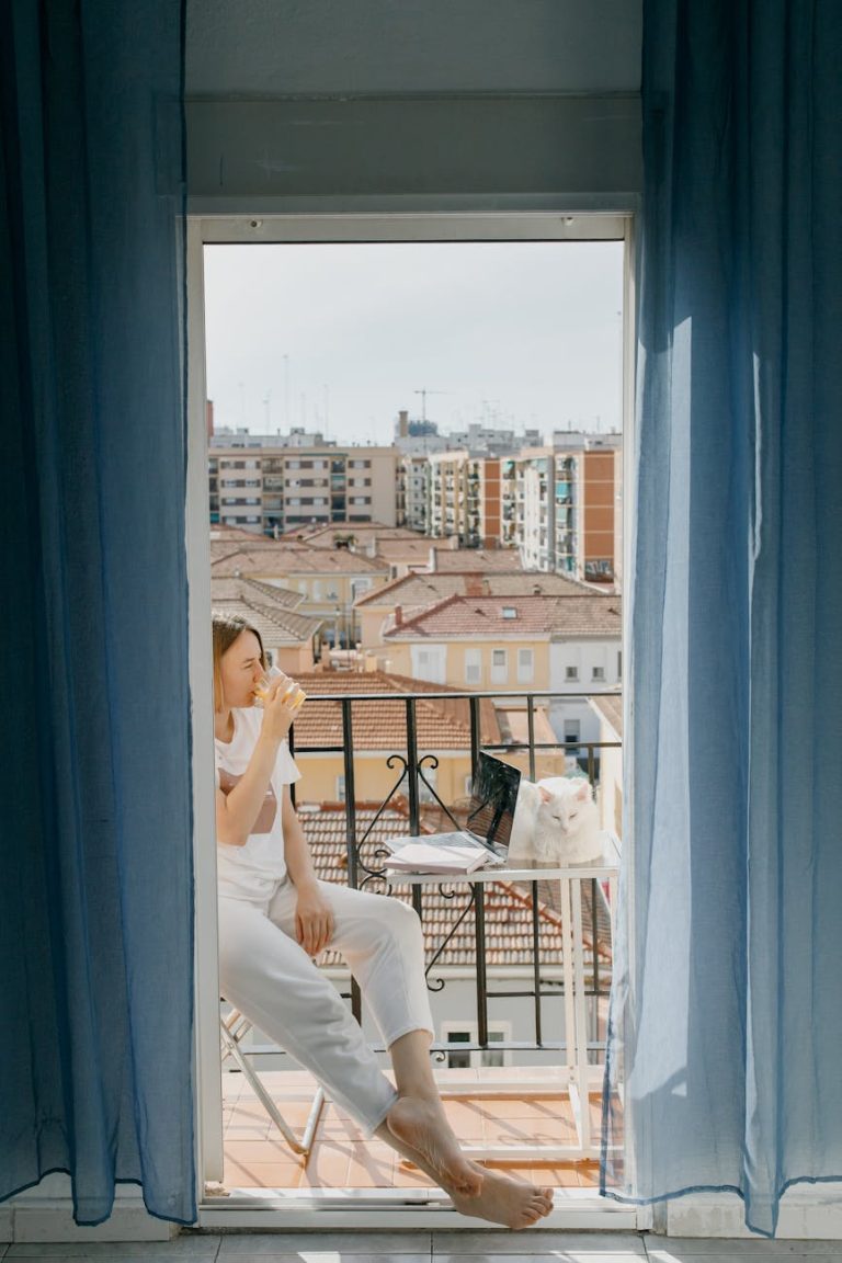 A young woman enjoys a quiet moment on her balcony, sipping coffee alongside her cat and laptop.