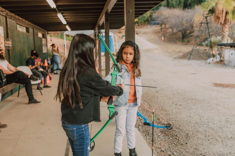 Two young girls practice archery at an outdoor range, learning to use a bow and arrow.