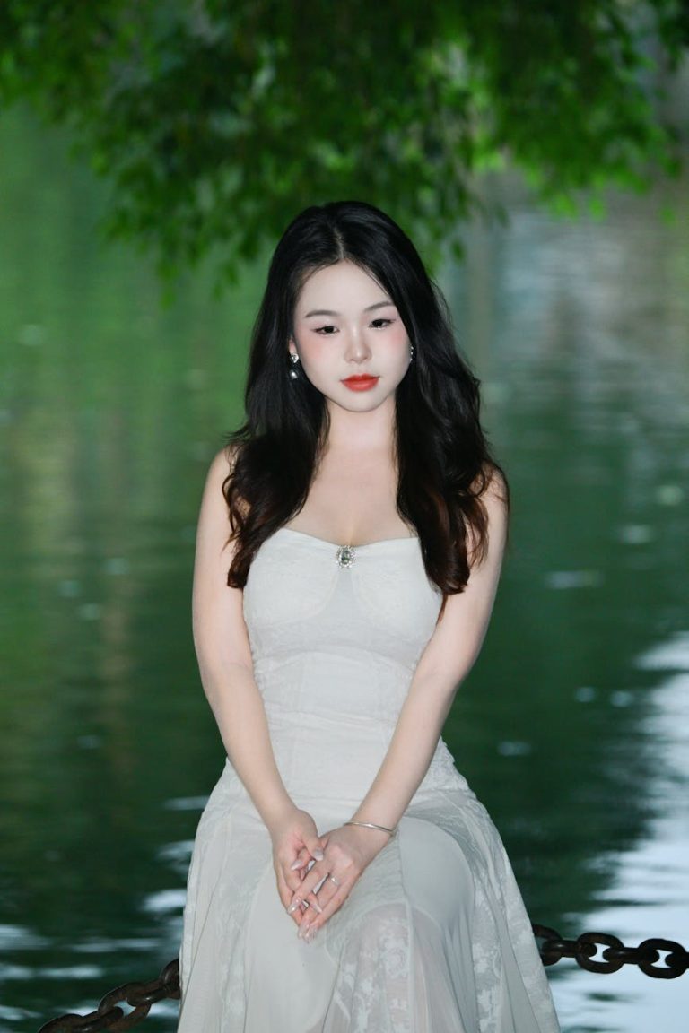 Portrait of a woman in a white dress posing by a serene lake, surrounded by greenery.