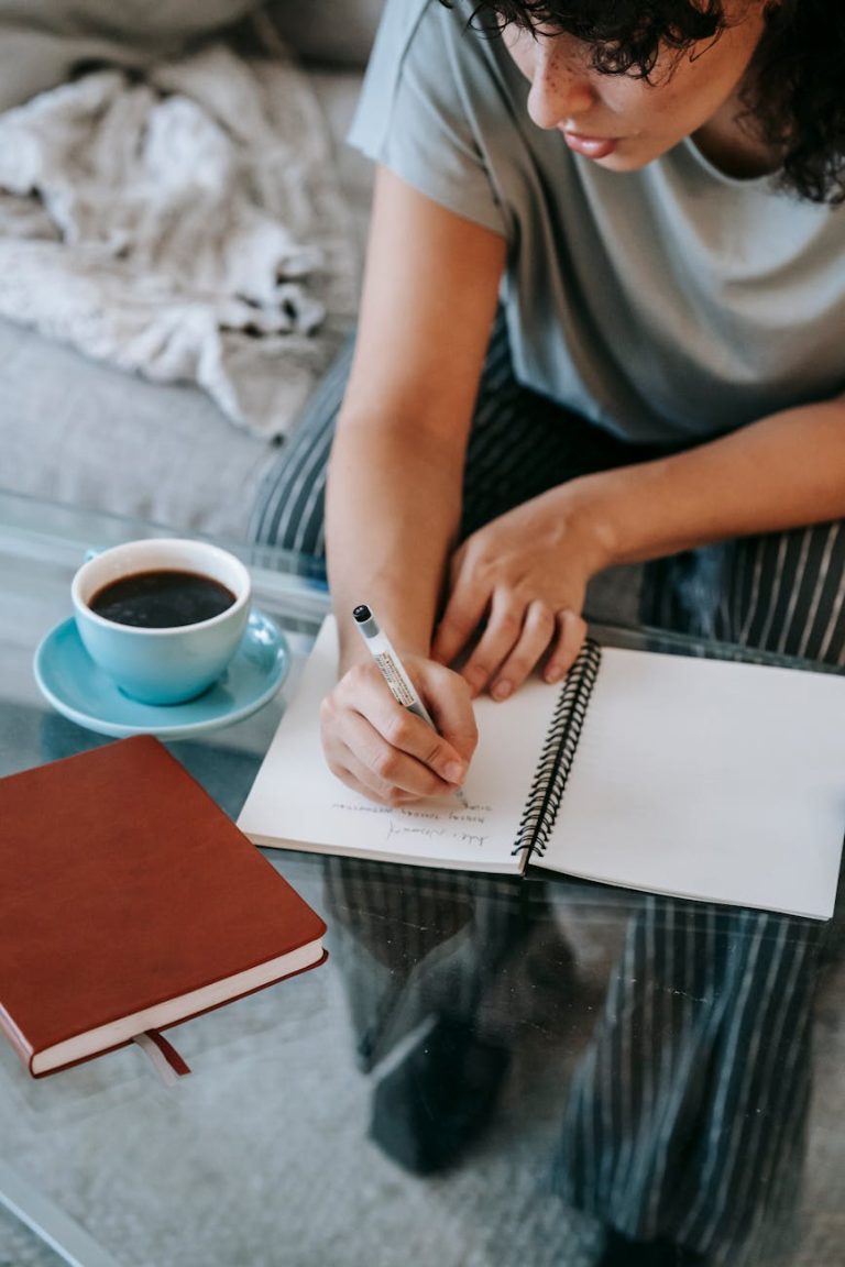 From above of crop young female student in casual clothes taking notes in copybook and drinking coffee while sitting on couch and doing homework assignment
