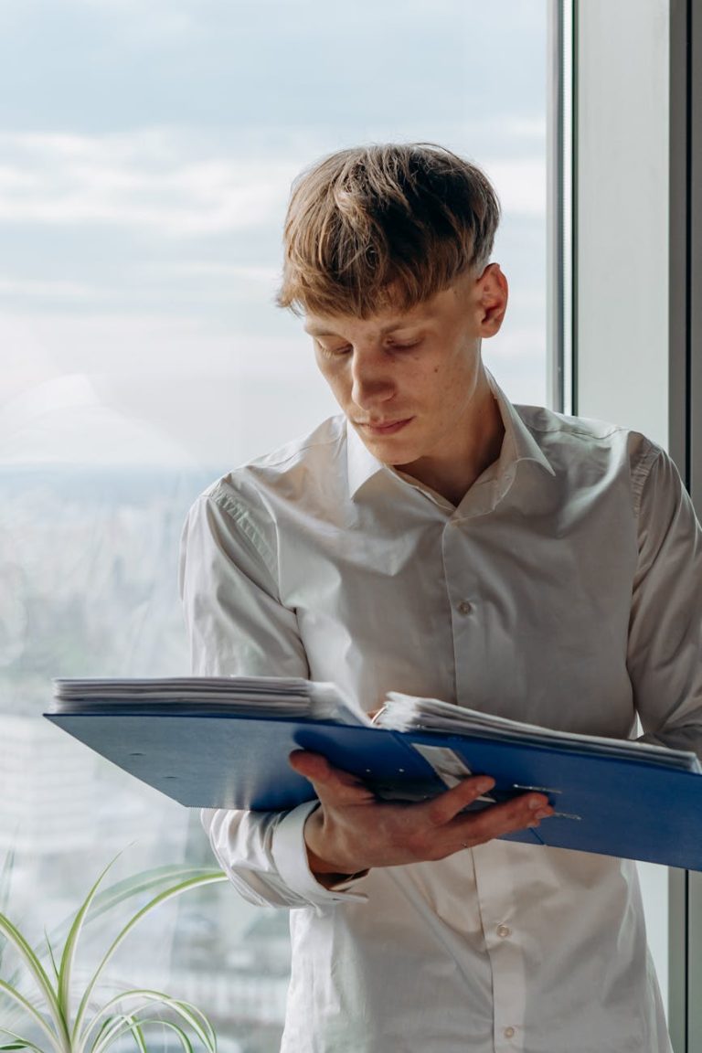Young man in white shirt reading binder by a window in an office setting.