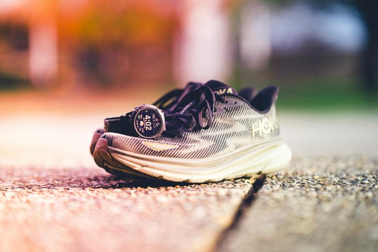 A pair of Hoka running shoes with a Garmin watch beside them on a sunny outdoor track.