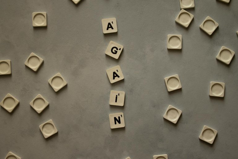 Top view of alphabet tiles spelling 'Again' on a neutral gray surface, surrounded by empty tile holders.